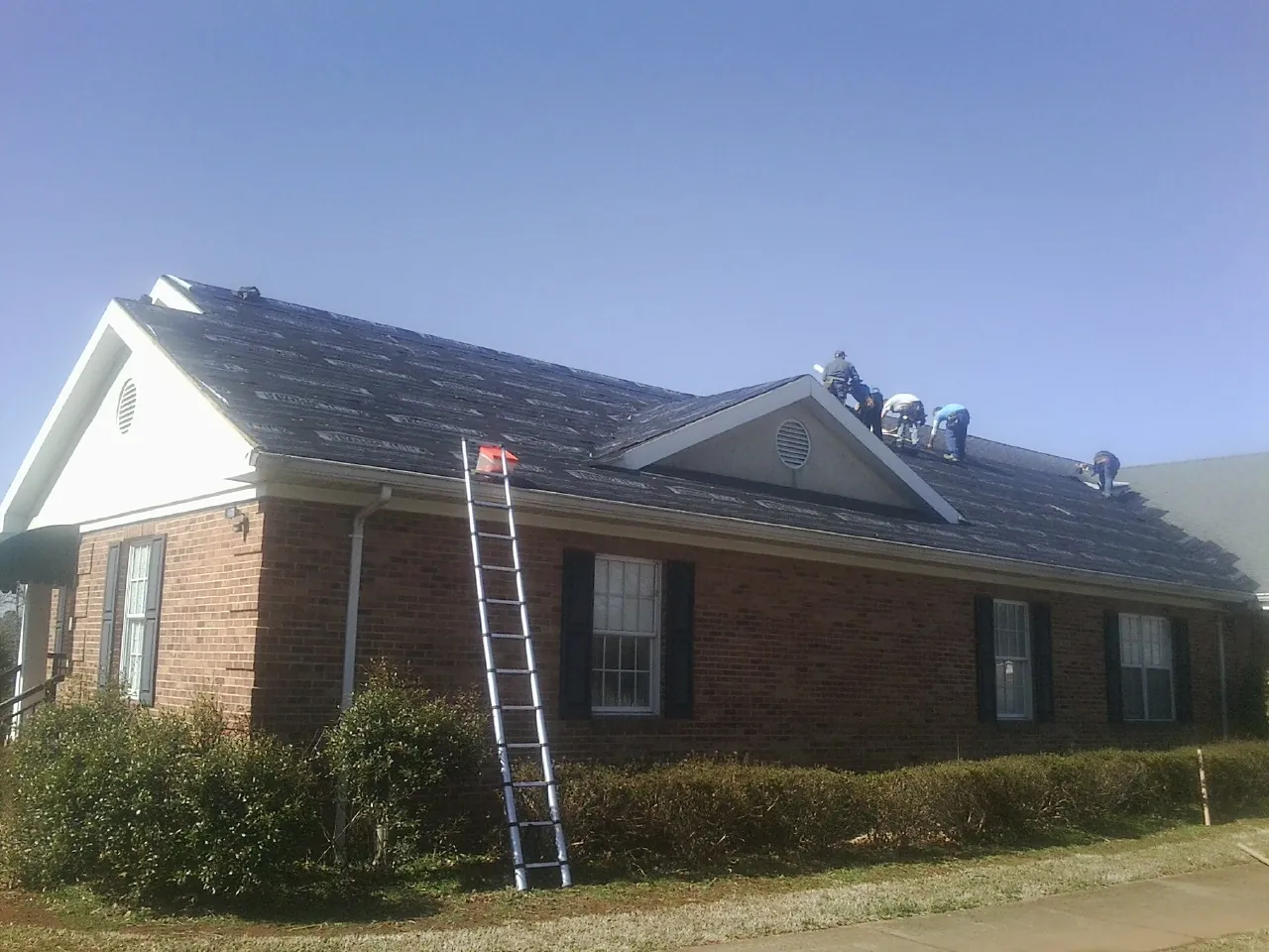 Crew tearing shingles off a brick ranch roof with a ladder set — Metal Roof Repair work by our crew in Westbury