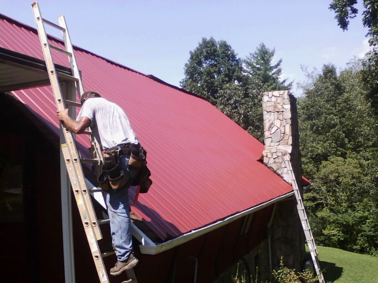 Roofer on a ladder beside a red metal roof and stone chimney — Soffit & Fascia Repair by Roofing Brothers in Westbury
